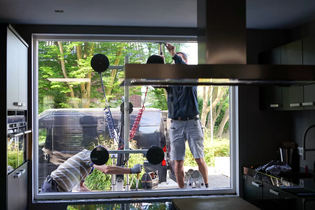 Workers installing a large glass window from outside a modern kitchen, using suction cups and tools.